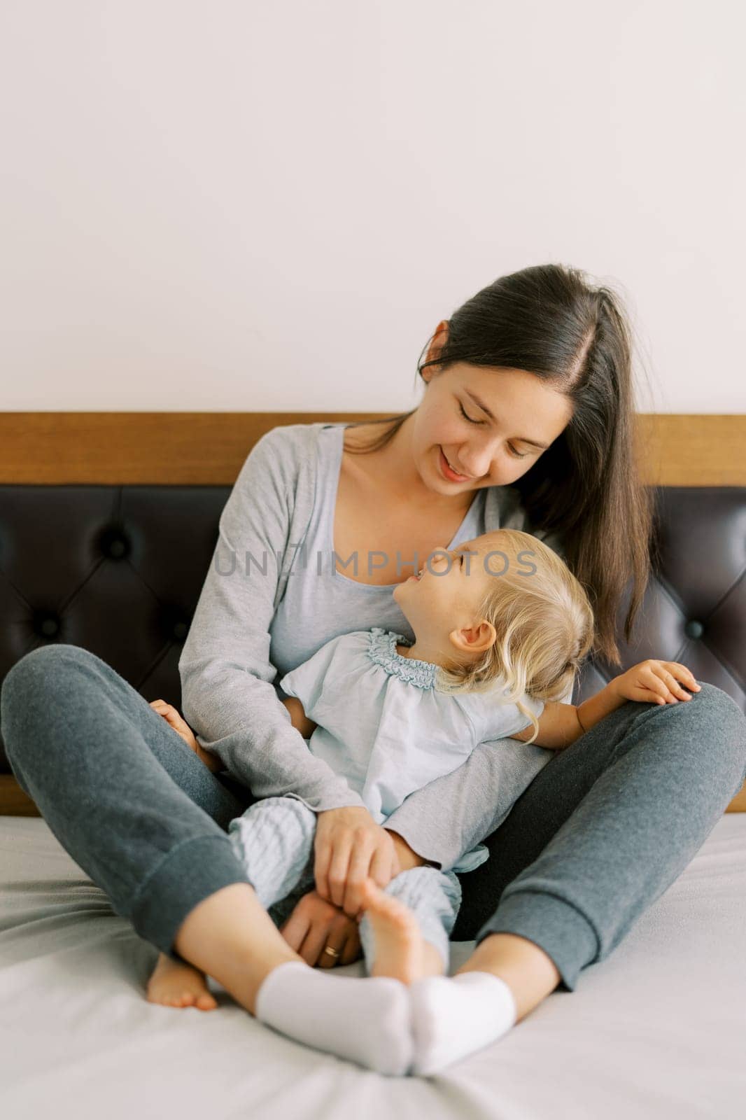 Little girl looks into the face of her mother hugging her from behind while sitting on the bed. High quality photo