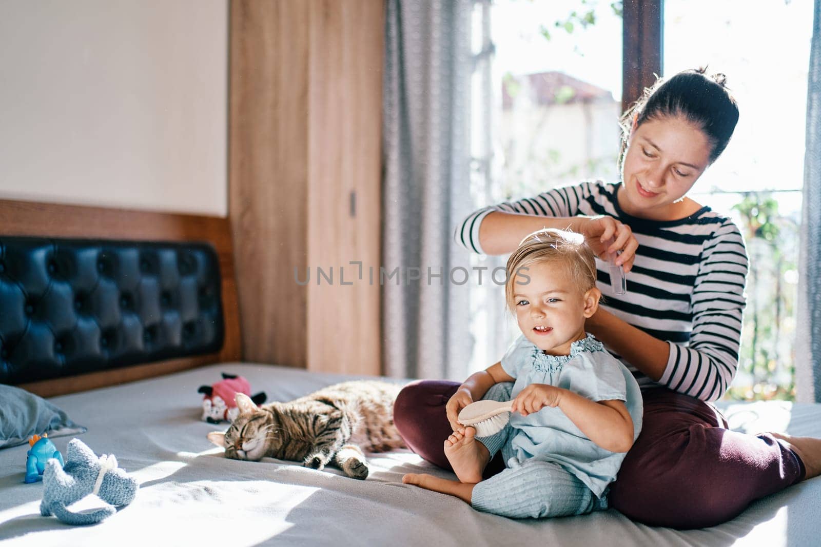Mom combs hair little smiling girl with a comb while sitting on the bed. High quality photo