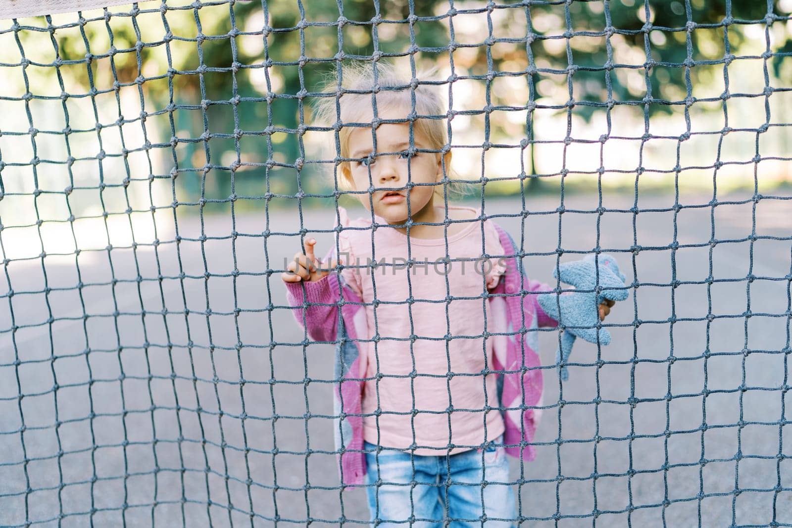 Little girl peeks through the cells of the rope net holding onto it with her fingers by Nadtochiy