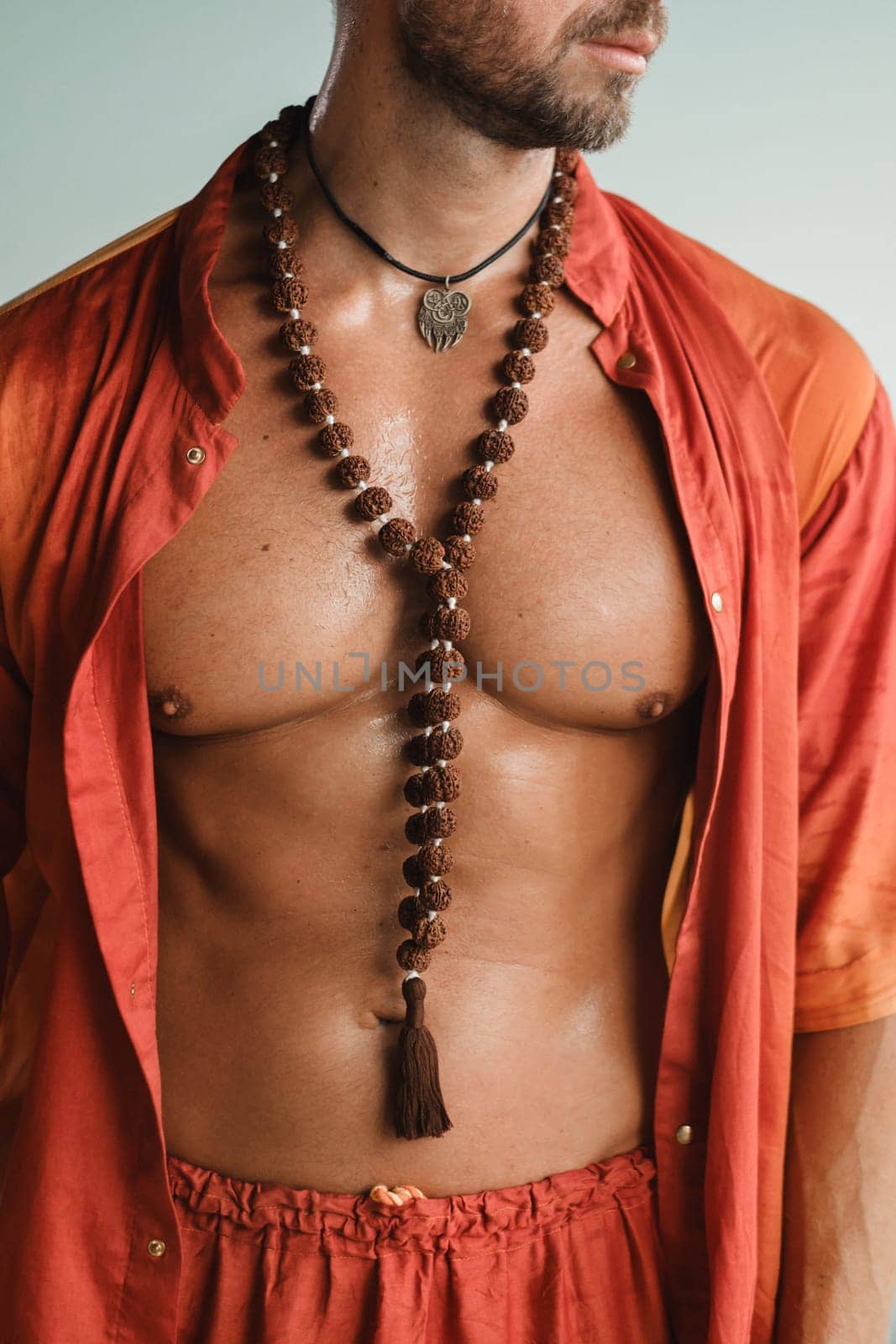 A muscular man with a naked torso in orange clothes stands in a room on a light background.