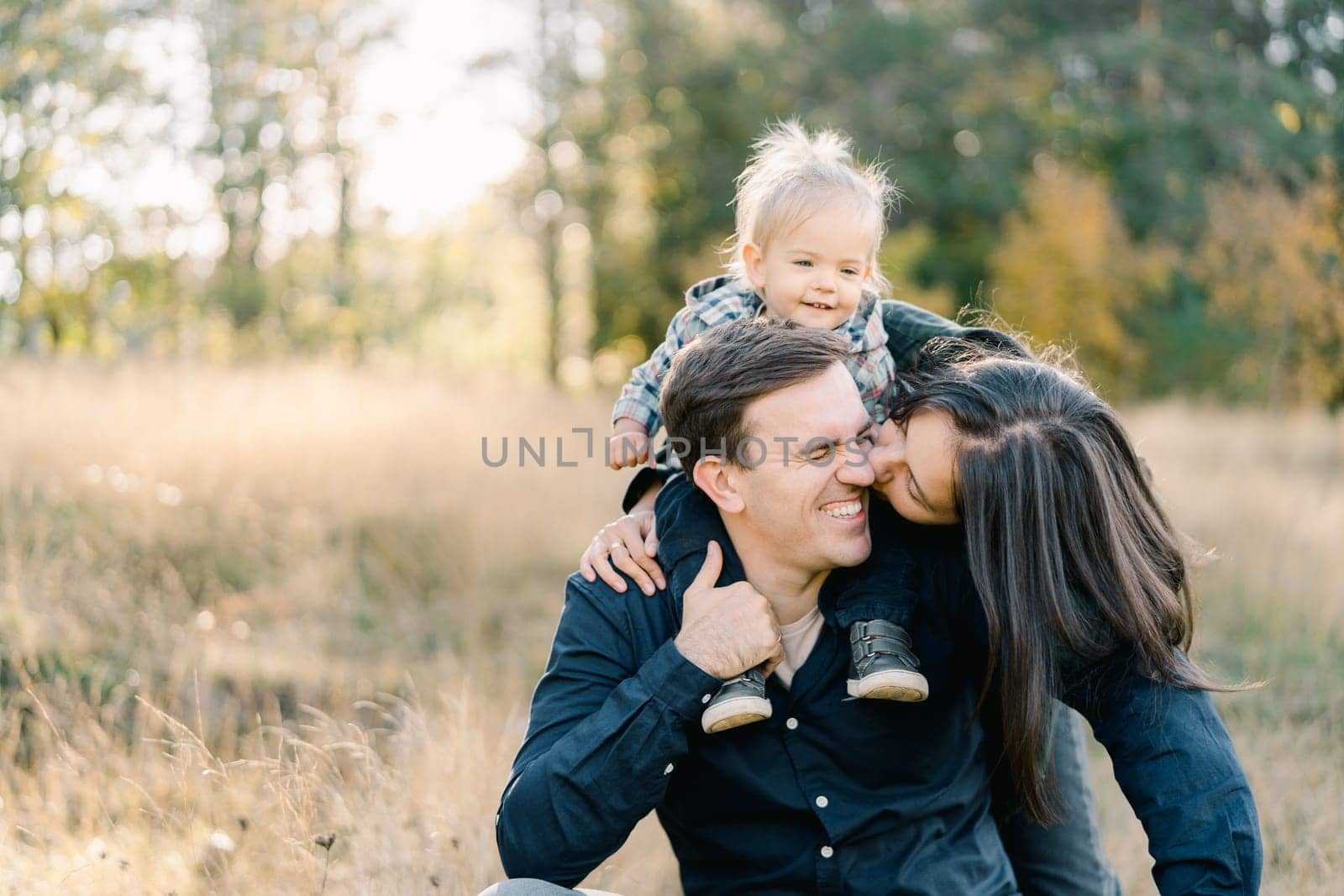 Mom kisses a laughing dad, from behind on the cheek with a little girl on his shoulders. High quality photo