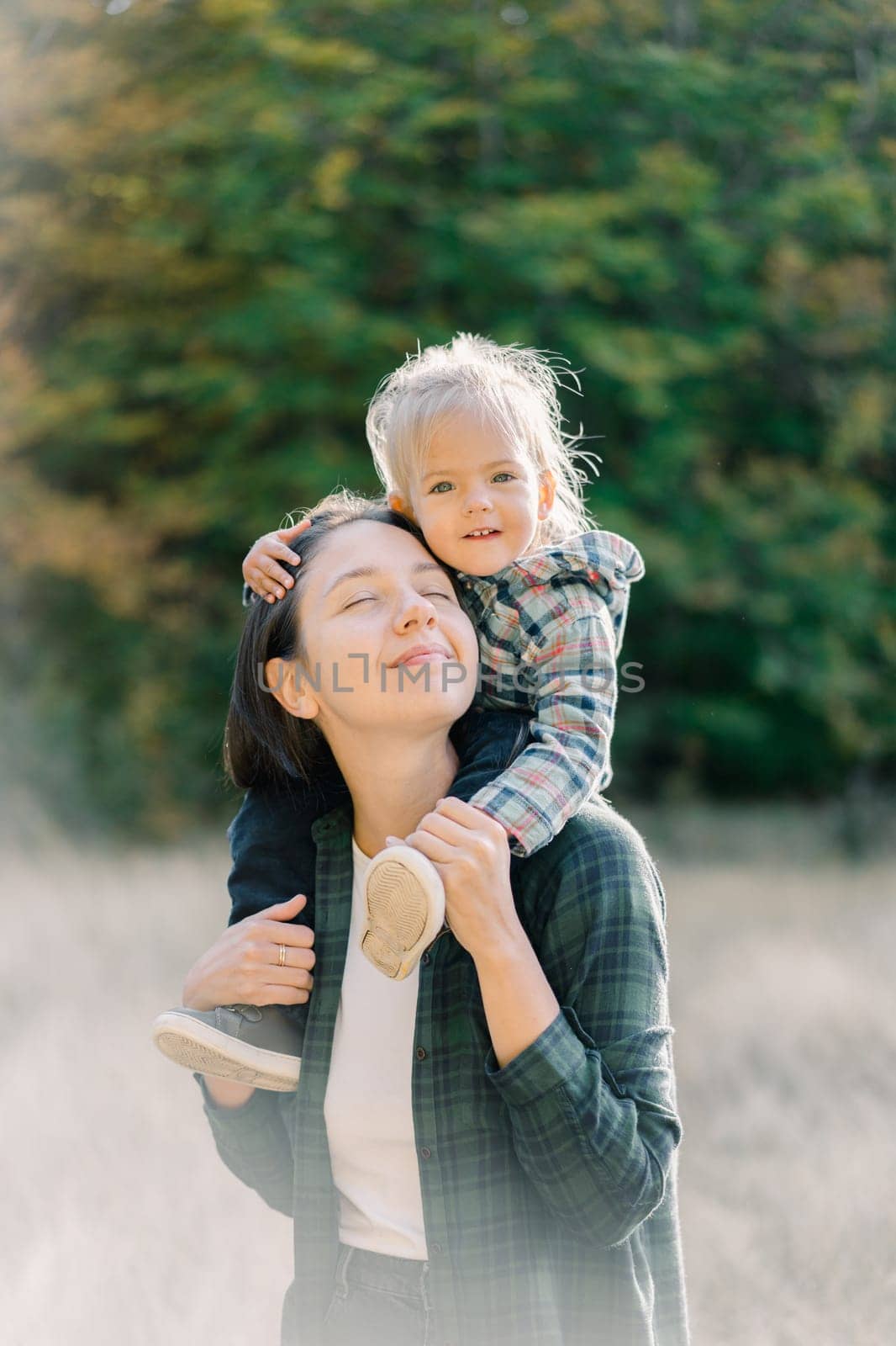 Smiling little girl hugging her mother head while sitting on her shoulders. High quality photo