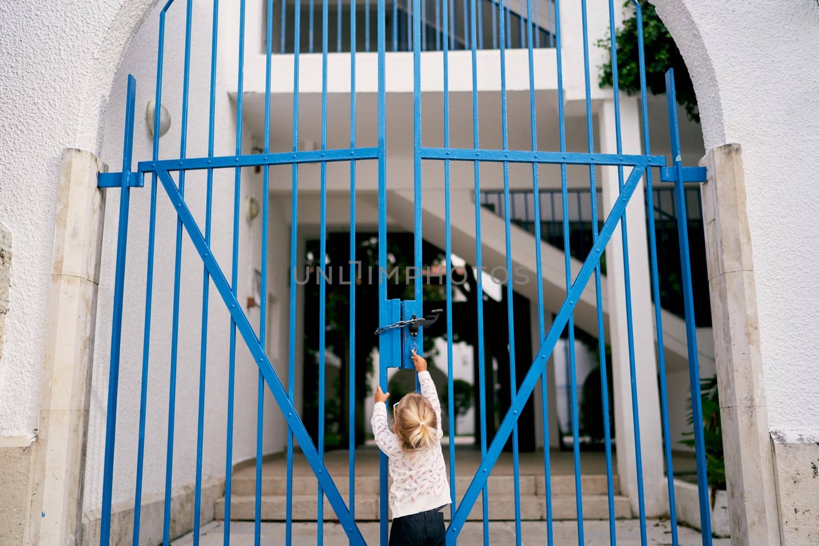 Little girl is trying to open the closed metal gate of the house while standing in the yard. Back view by Nadtochiy