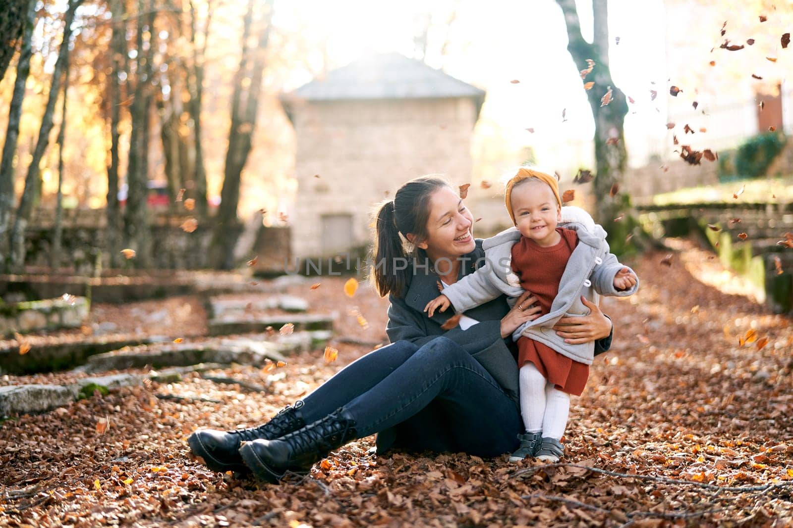 Mom sits on the ground hugging a little girl standing nearby under the falling autumn leaves. High quality photo