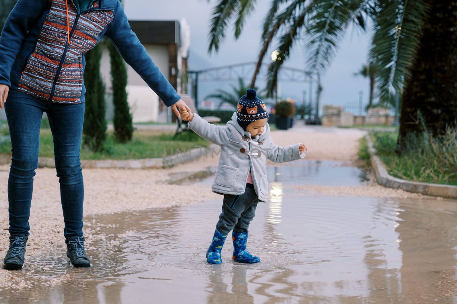 Little girl in rubber boots stands in a puddle holding her mother hand. Cropped by Nadtochiy