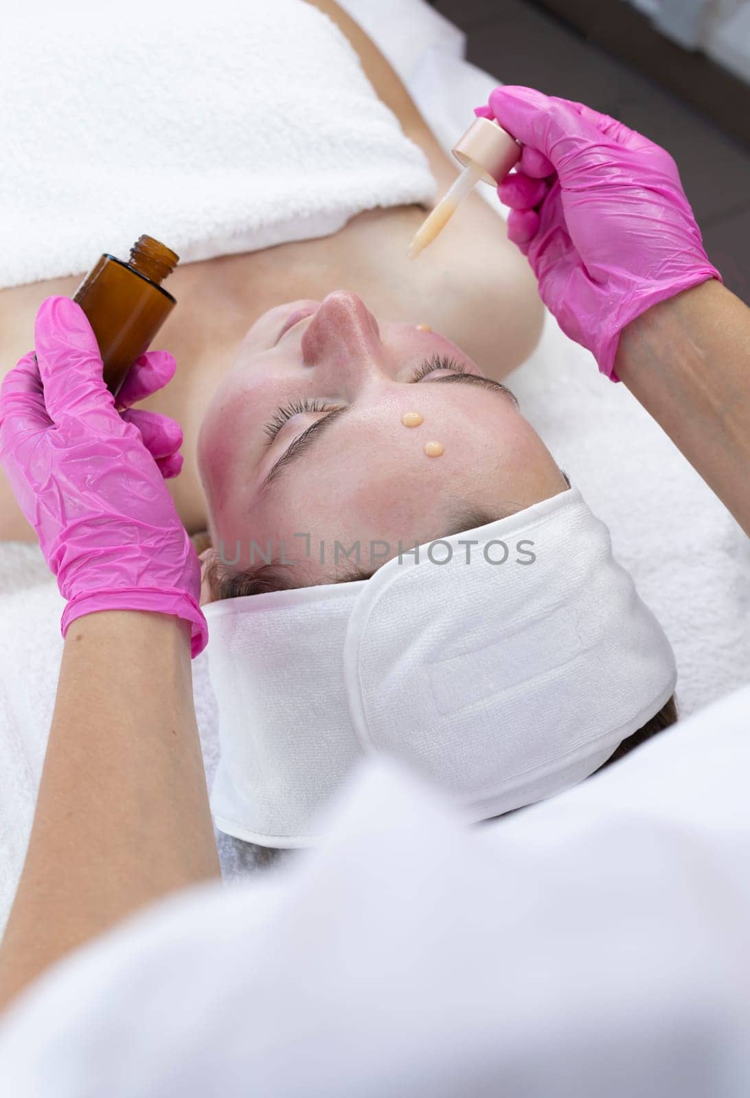 Beauty Specialist Applies Serum To Patient's Face With Pipette After Peeling Procedure In Spa Salon. Facial Skin Care With Treatment Cosmetic Oil And Serum. Woman Enjoys Skin Moisturizing Procedure.