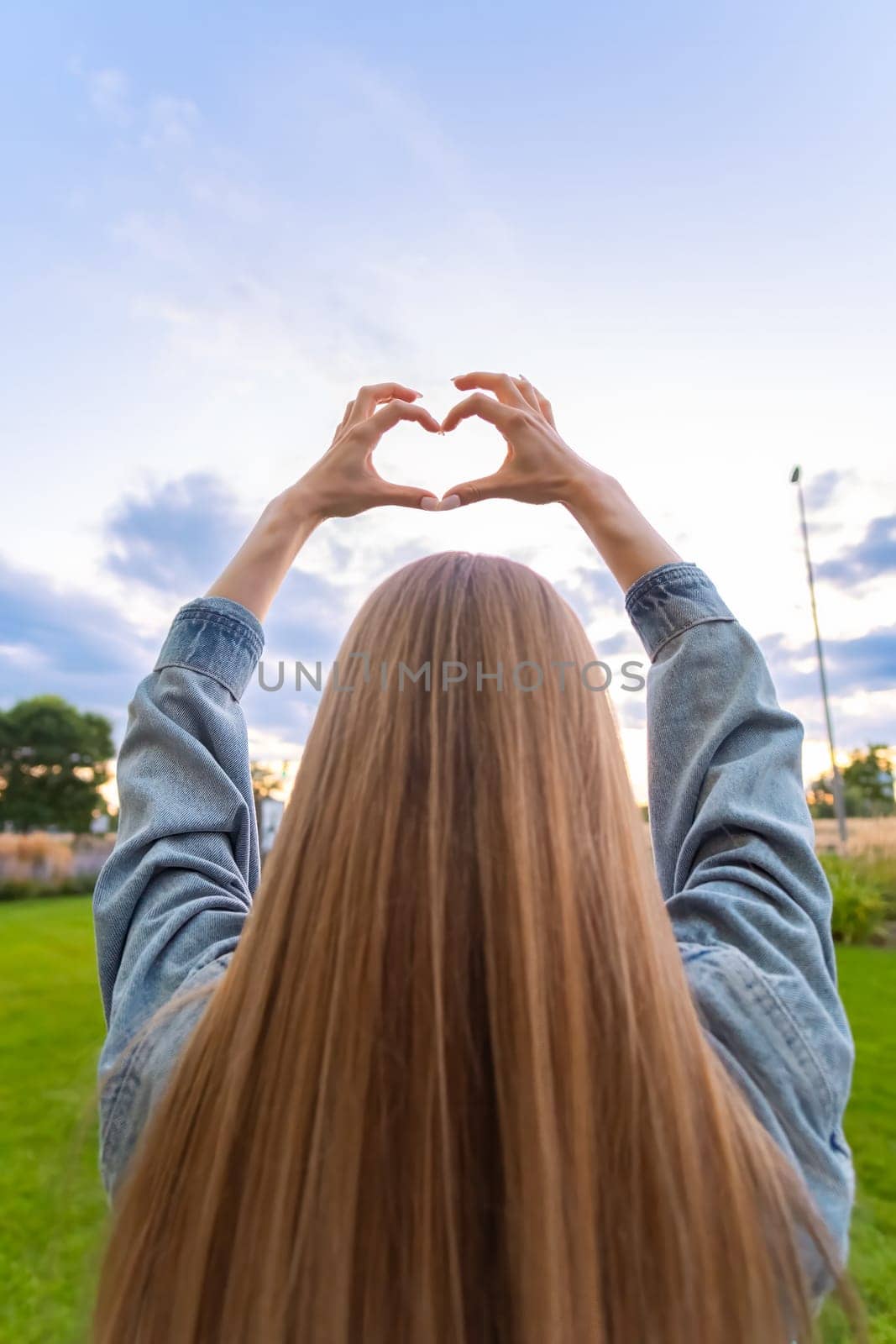 Young blonde woman with long hair making hearts with her hands against sky at sunset by vladimka