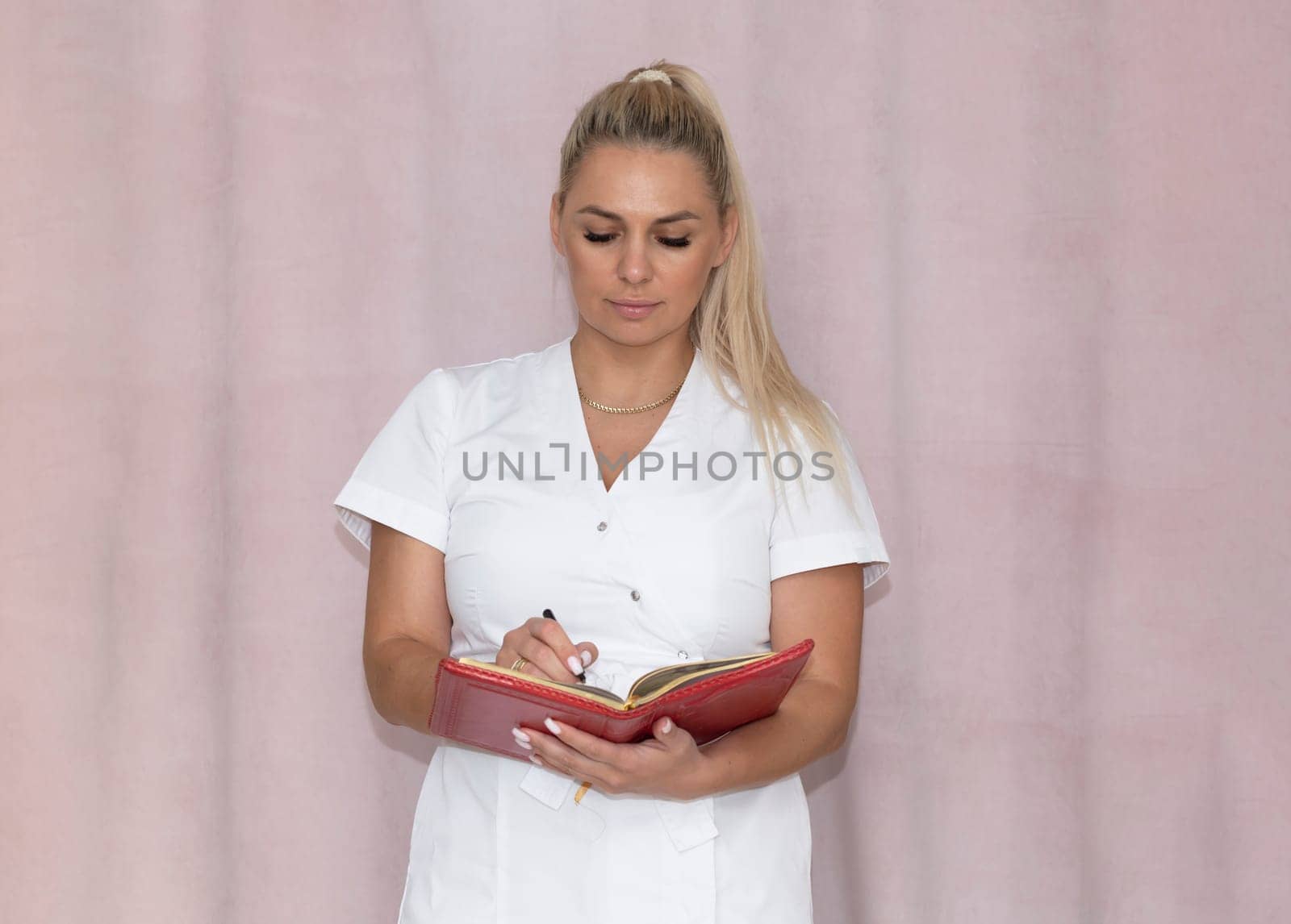 Real Beautiful Aesthetician, Beauty Doctor in White Scrubs holds a Book and Makes Notes on Pink Background in Salon. Medical Beauty Procedure Appointment or Consultation. Horizontal Plane.