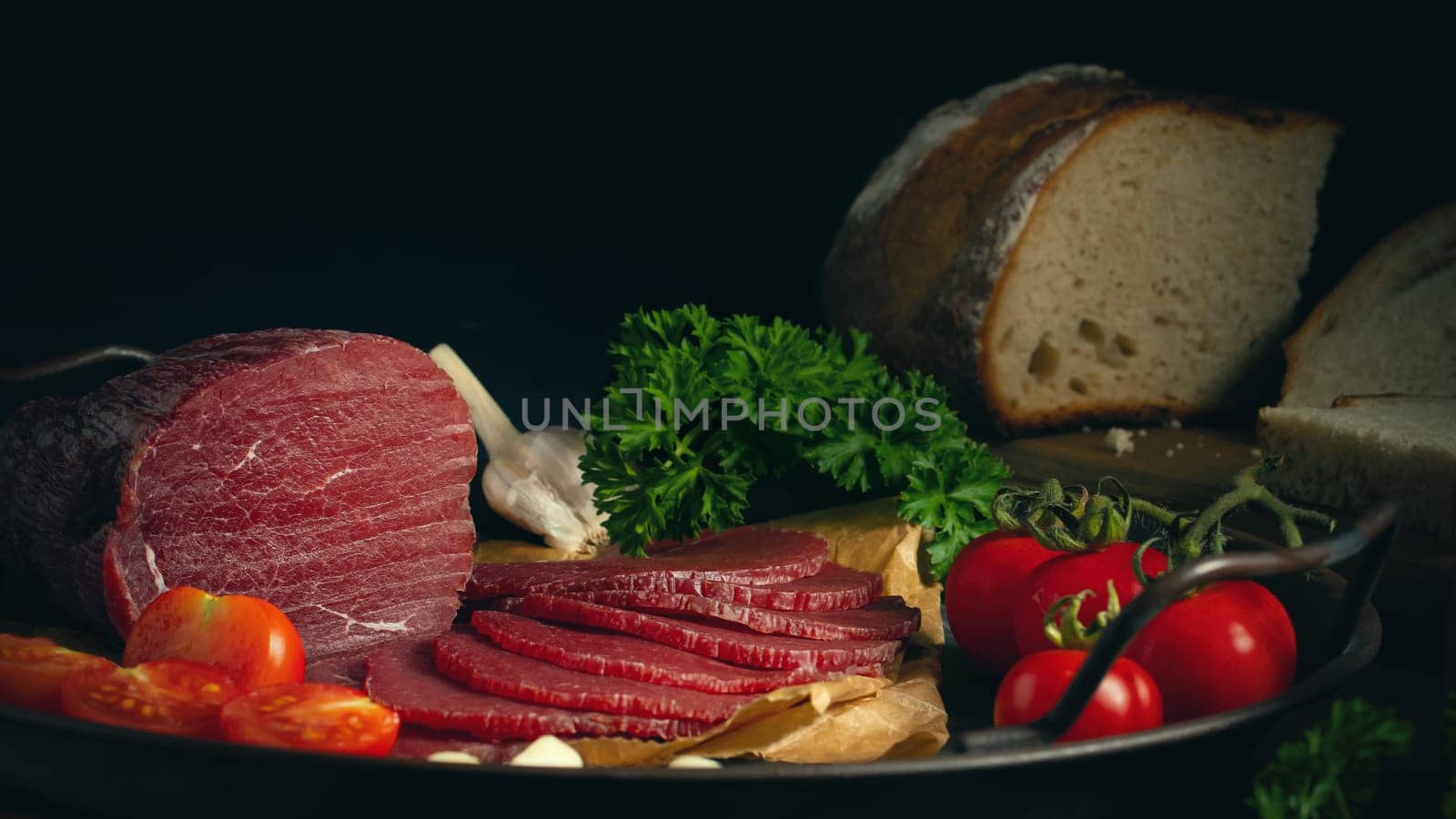 Whole and sliced bresaola on a metal round tray with tomatoes, garlic and bread.