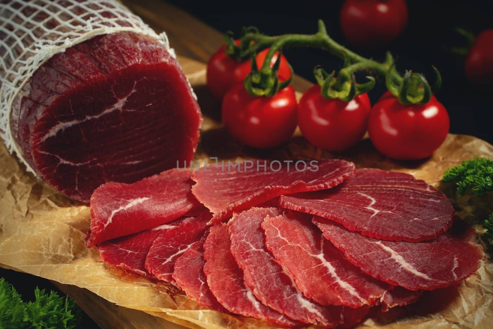 Whole and sliced bresaola on paper on a cutting board with tomatoes.