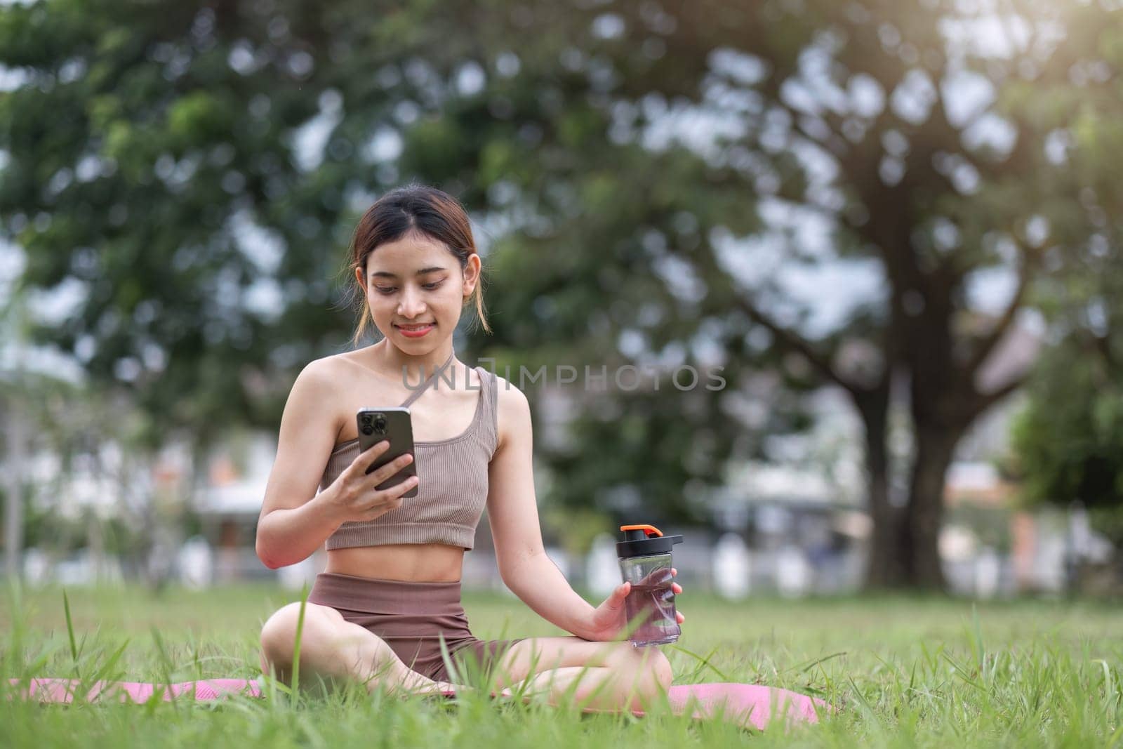 Young woman sitting on an exercise mat Use a smartphone during an outdoor yoga activity on the grass at a park..