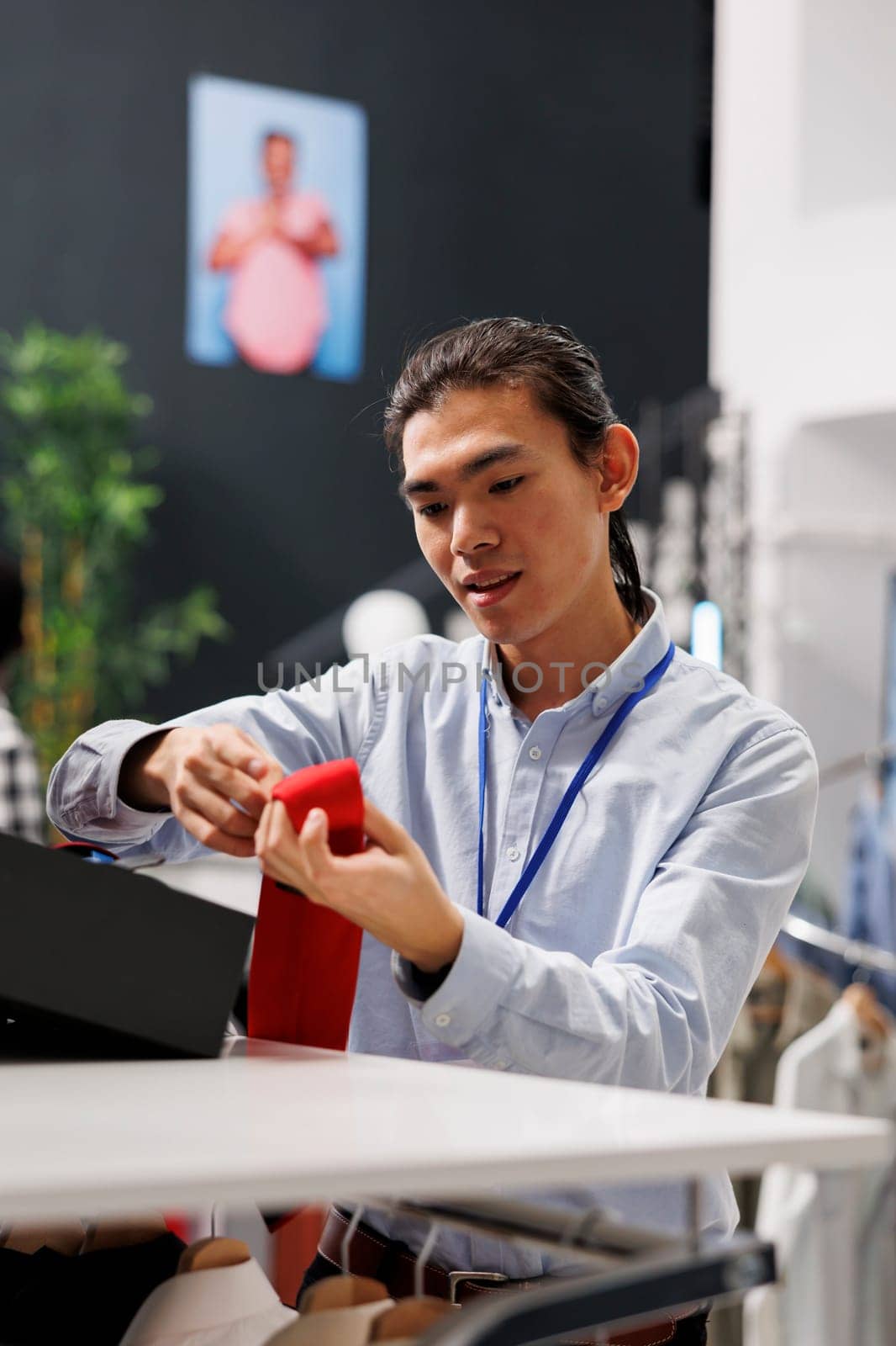 Asian man looking at fashionable accessories, arranging belt on store shelf in modern boutique. Stylish employee working in shopping centre, working with hangers full with sales clothes