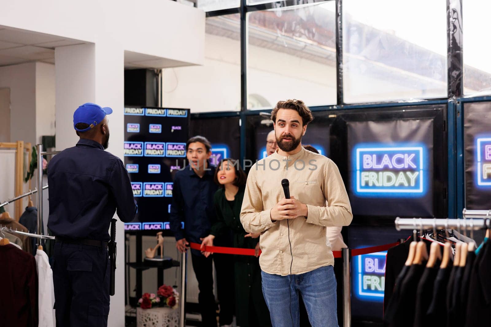 Young man TV reporter covering news real-time, live streaming during Black Friday sales. Correspondent standing at store entrance talking into microphone making live newscast about shopping madness