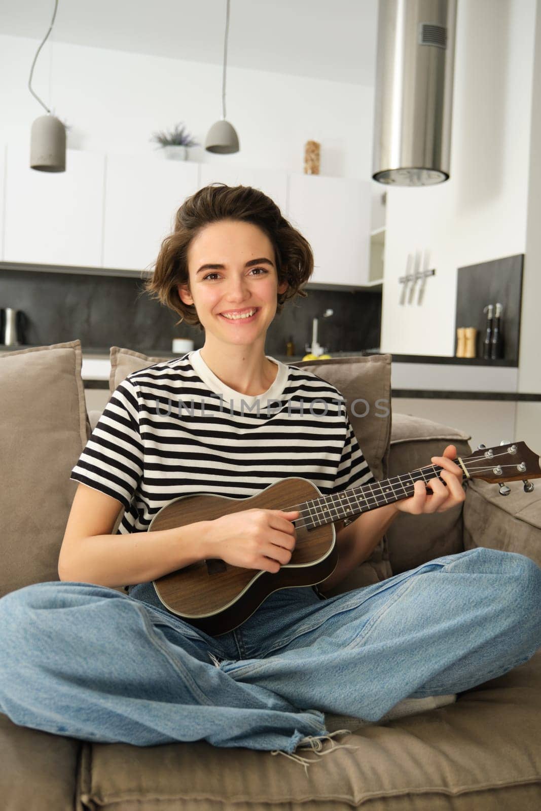 Vertical shot of young musician, woman learns how to play ukulele, sits on sofa with crossed legs and smiling at camera.