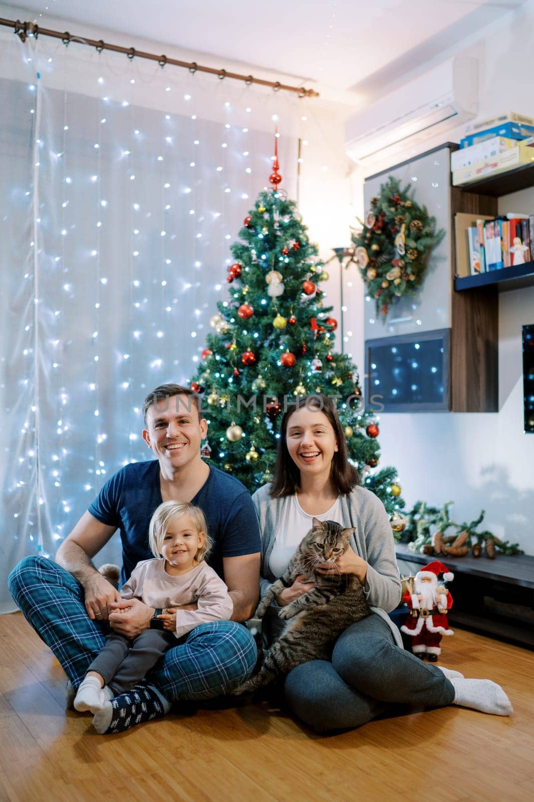 Happy parents with a little girl and a cat sit near the Christmas tree on the floor. High quality photo