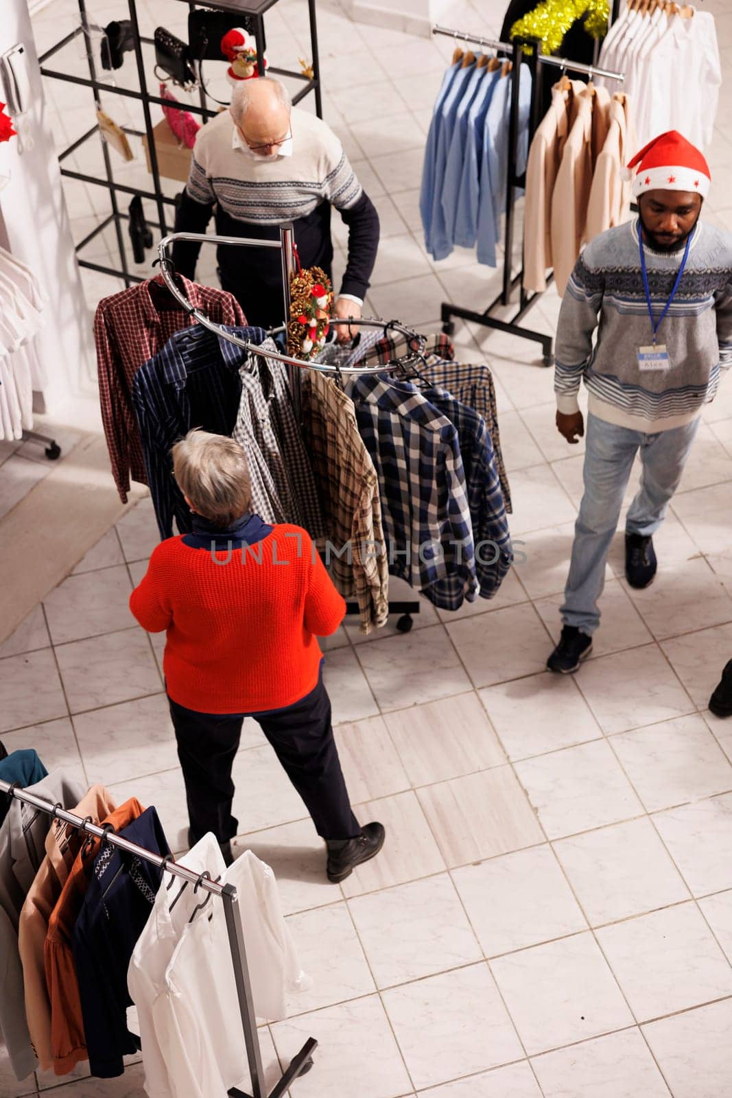 Various shoppers looking at clothing items on racks in shopping center while seeking for garments to purchase. Consumers going apparel shopping as gifts for festive season, Christmas eve.