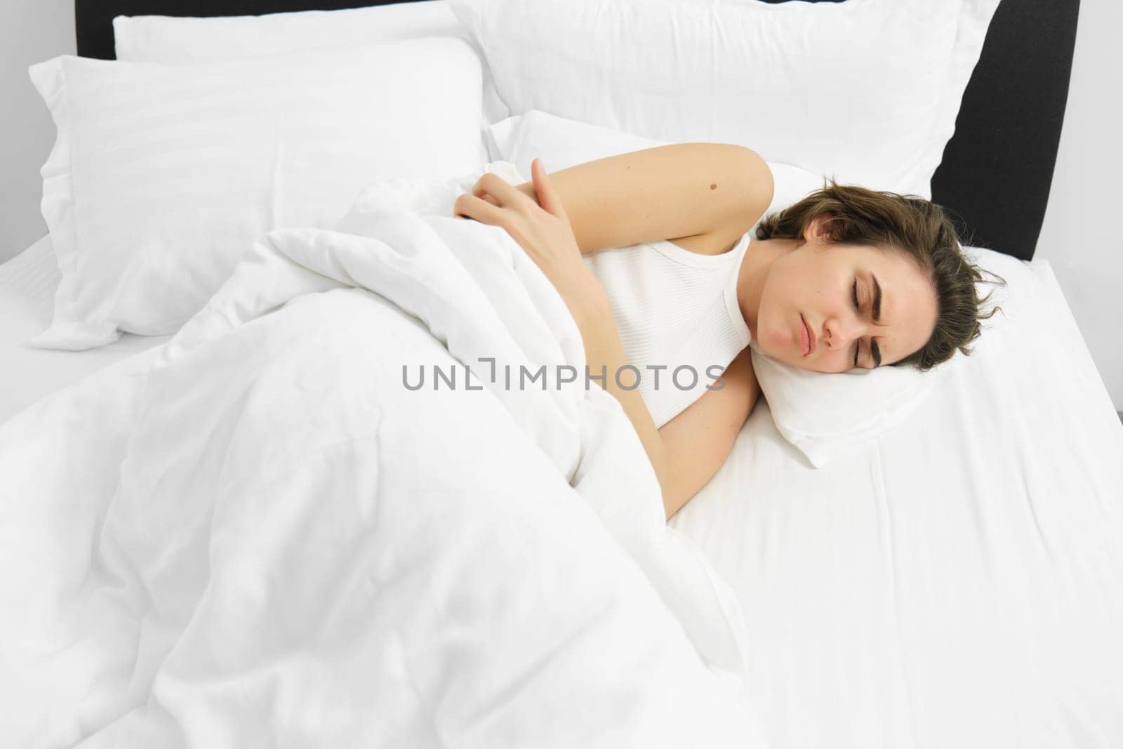Portrait of woman lying in bed with stomach ache, has menstrual cramps, frowning and feeling pain period by Benzoix