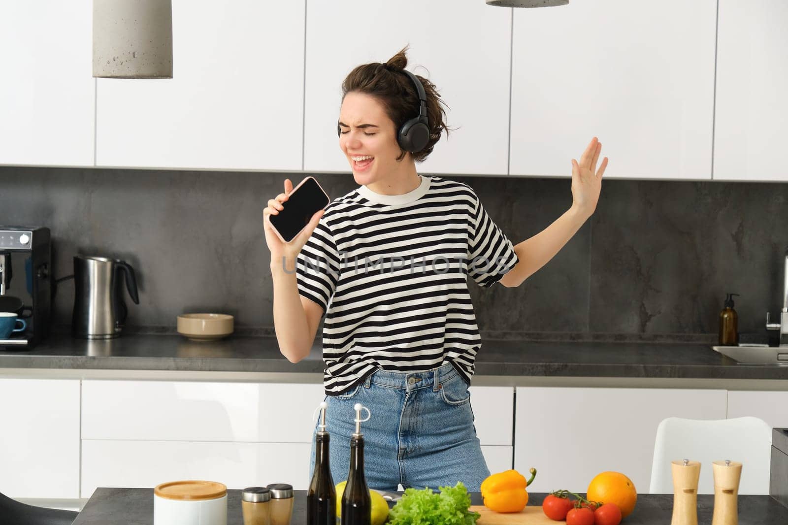 Portrait of happy dancing woman, cooking in the kitchen and listening music in headphones, chopping vegetables for salad and singing with carefree face.