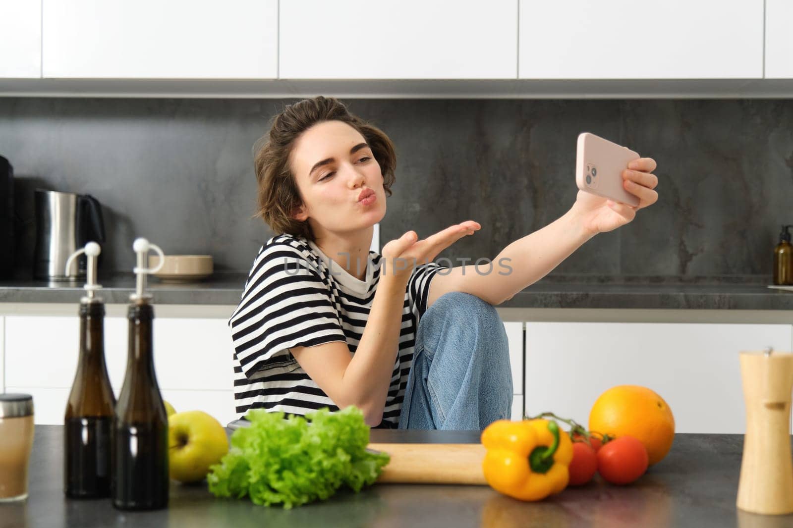 Cute young woman, lifestyle blogger cooking, sitting in the kitchen with vegetables and chopping board, taking selfie on her new smartphone, sending kisses at mobile camera by Benzoix
