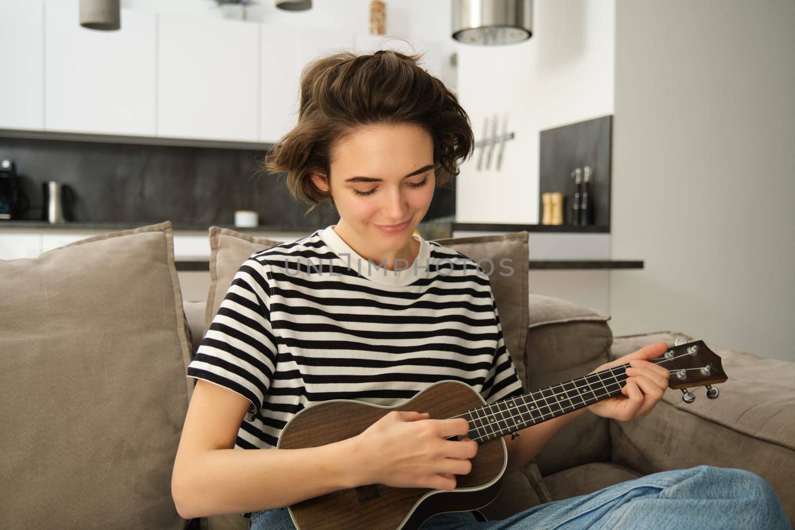Portrait of cheerful young woman playing her ukulele, singing and laughing, sitting in living room at home. Lifestyle and music concept