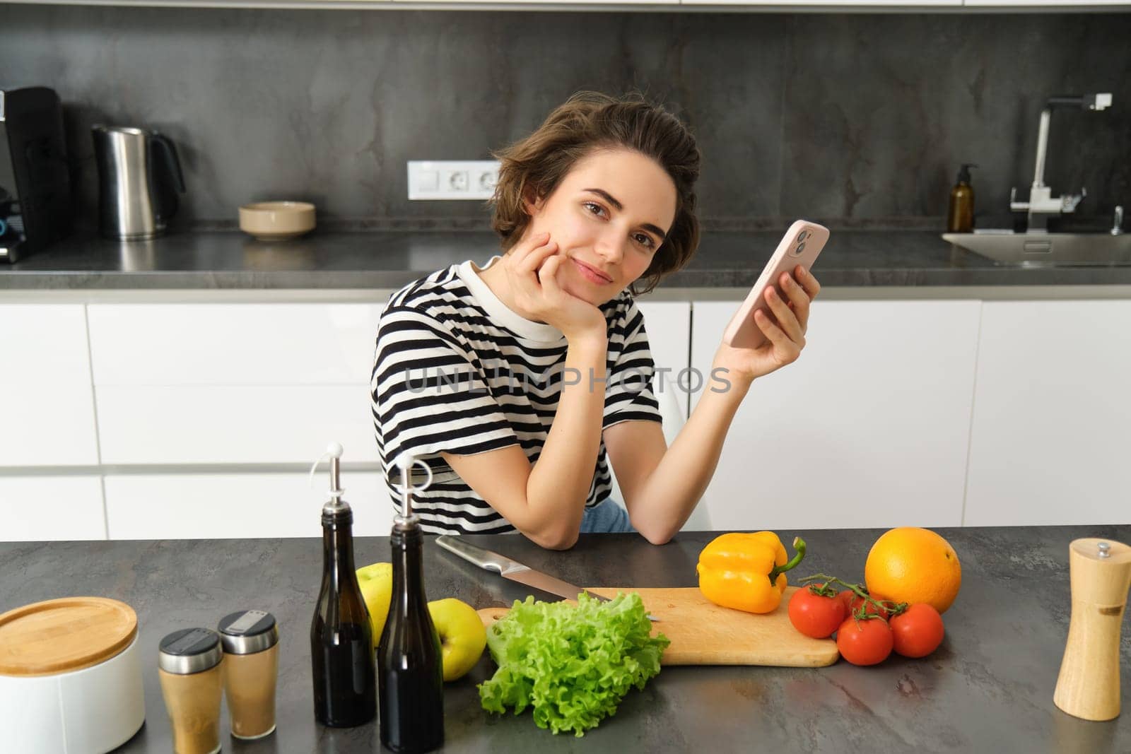 Portrait of cute brunette woman in the kitchen, cooking, searching recipe on smartphone, taking pictures for her food blog, cooking meal from vegetables, vegan salad by Benzoix