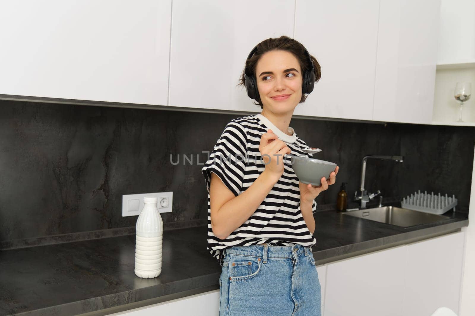 Portrait of modern woman eats breakfast, holds bowl and spoon, stands in kitchen, listens music in wireless headphones. Concept of lifestyle and home by Benzoix