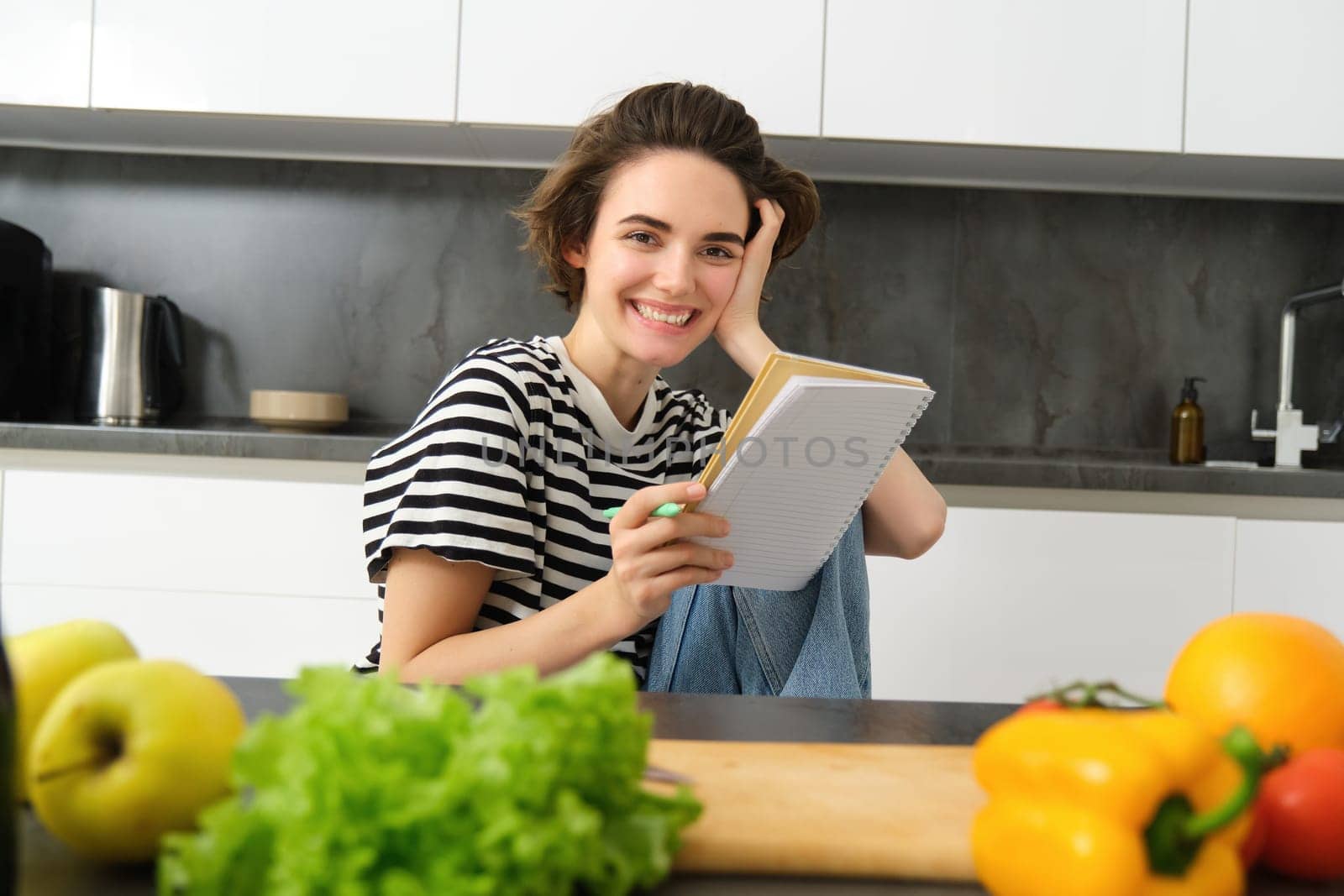 Portrait of young smiling woman in kitchen, holding notebook, making notes for recipe, writing grocery list, cooking salad, sitting near vegetables and chopping board by Benzoix