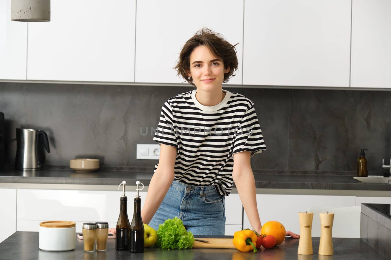 Portrait of beautiful young woman, female model posing near chopping board with vegetables and salad dressing, making herself light diet meal, vegan food for breakfast, standing in kitchen by Benzoix