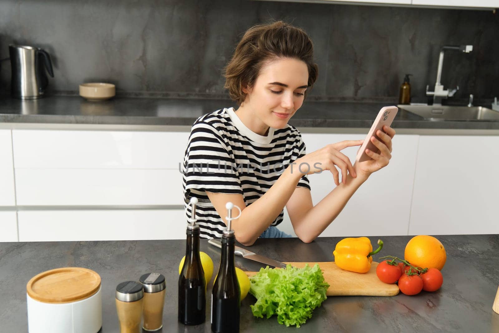 Portrait of young woman with smartphone, sitting in the kitchen and cooking salad, searching for healthy recipe online, has vegetables and chopping board on the table by Benzoix