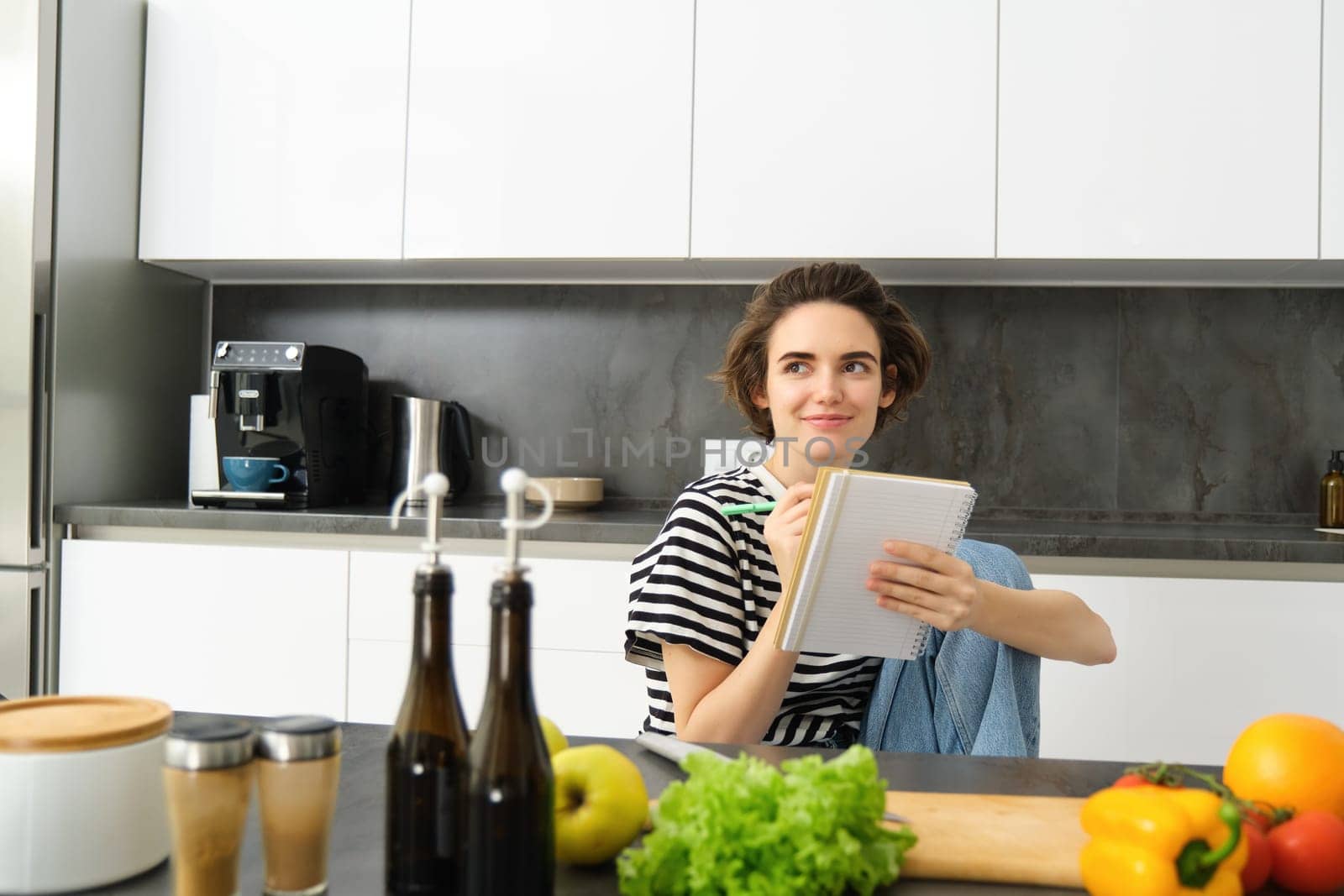 Portrait of young woman looking at cooking ingredients on kitchen counter and making notes, writing down recipes, thinking of meal for dinner, preparing vegetarian food.