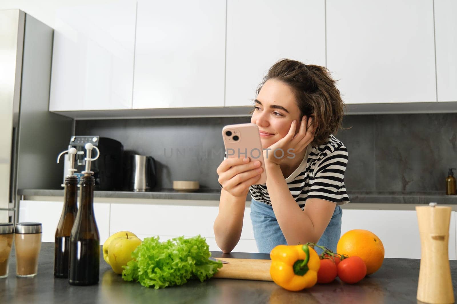 Portrait of young smiling woman, searching cooking recipe online on smartphone, standing near vegetables and chopping board, making meal, preparing salad and using social media on mobile phone by Benzoix