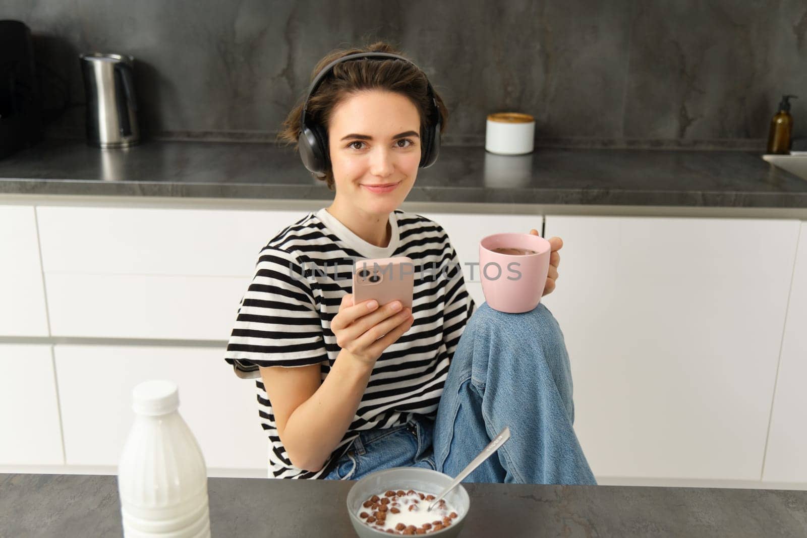 Modern female model smiling, using mobile phone, drinking tea and eating breakfast in kitchen. Lifestyle concept