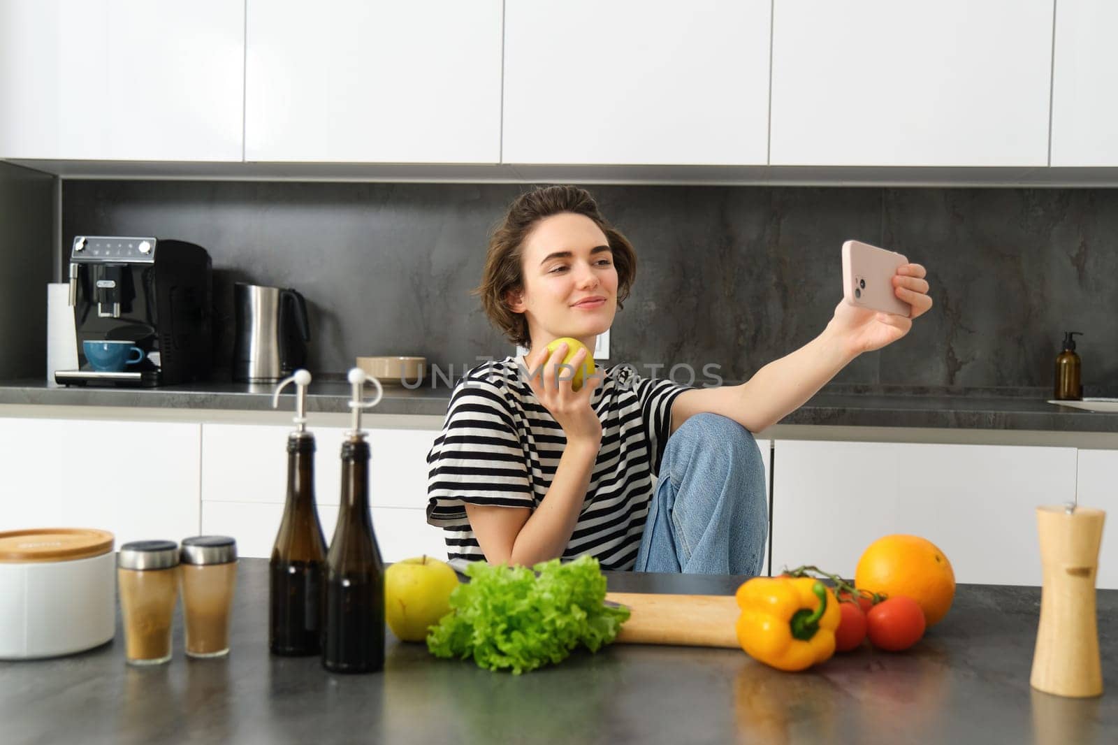 Portrait of beautiful smiling woman in the kitchen, taking selfie with apple, cooking healthy meal from vegetables, chopping food ingredients and recording video on mobile phone by Benzoix