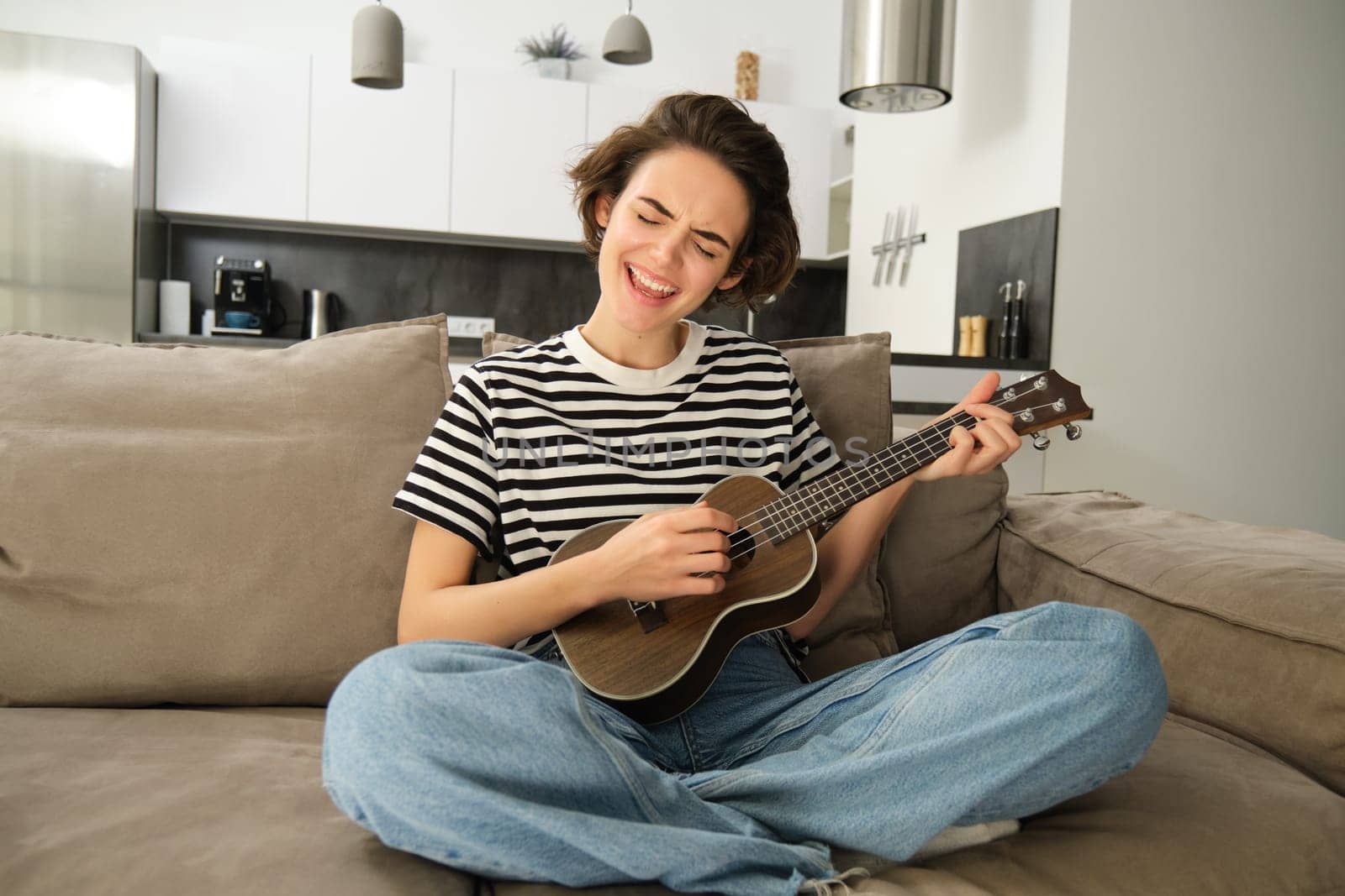 Portrait of cheerful young woman playing her ukulele, singing and laughing, sitting in living room at home by Benzoix