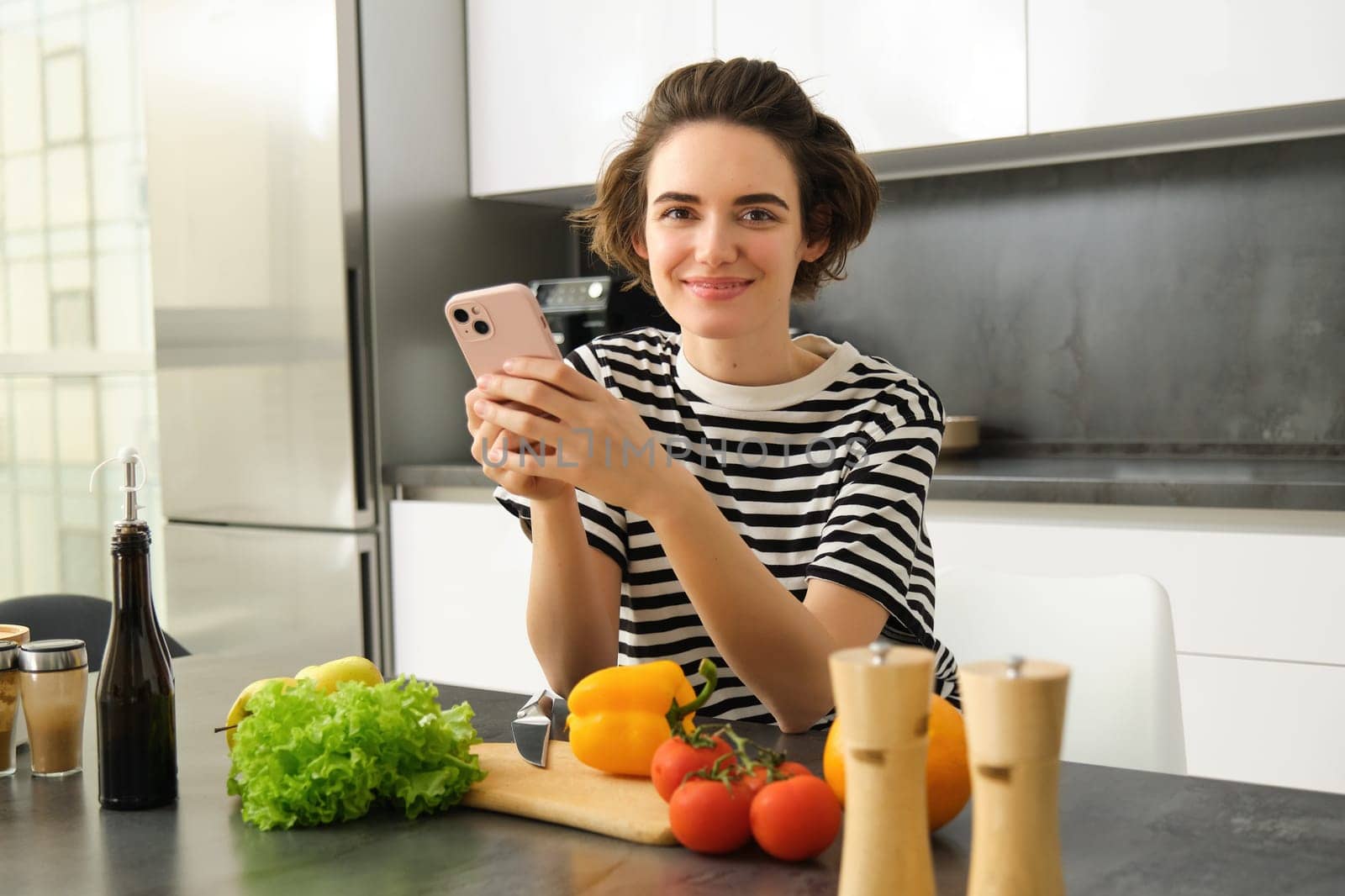 Portrait of young woman standing in the kitchen with vegetables and chopping board, holding smartphone, searching healthy diet recipes on mobile phone browser, cooking at home by Benzoix