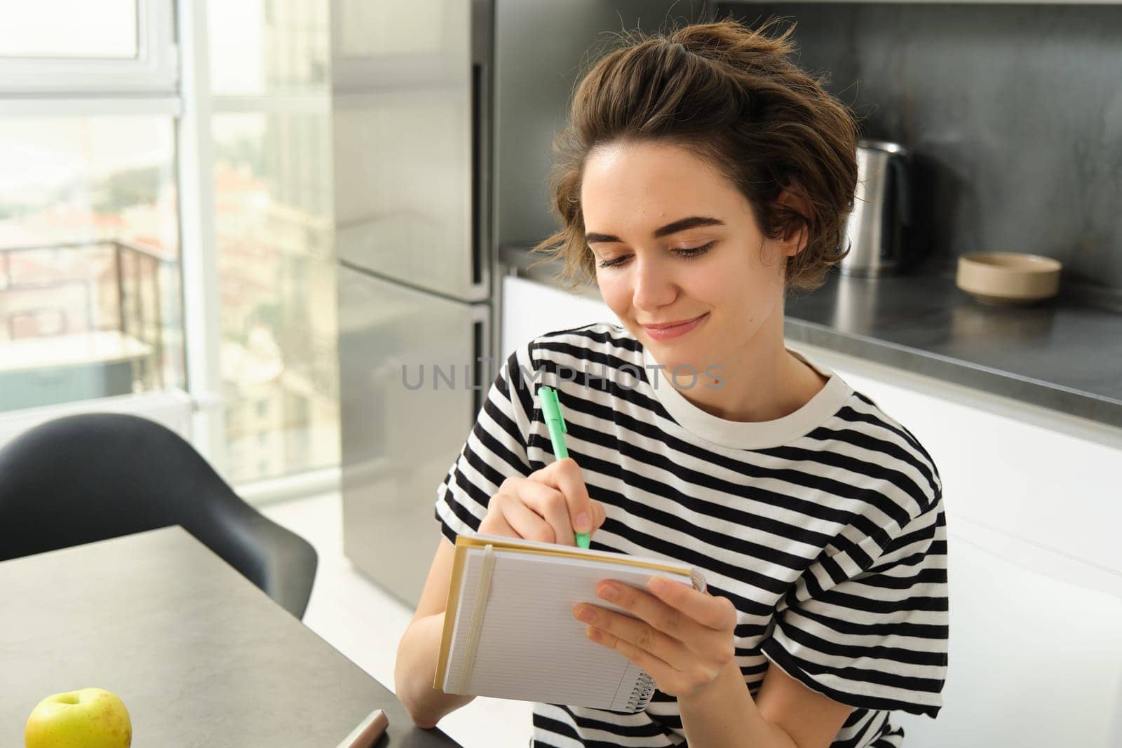 Close up portrait of smiling, cute woman with notebook, writing down recipe, making notes or grocery list for shopping, sitting in the kitchen.