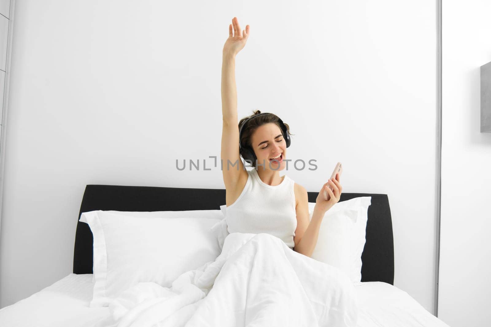 Portrait of excited, happy woman dancing and singing in bed, waking up and listening to music in wireless headphones. Lifestyle and wellbeing concept