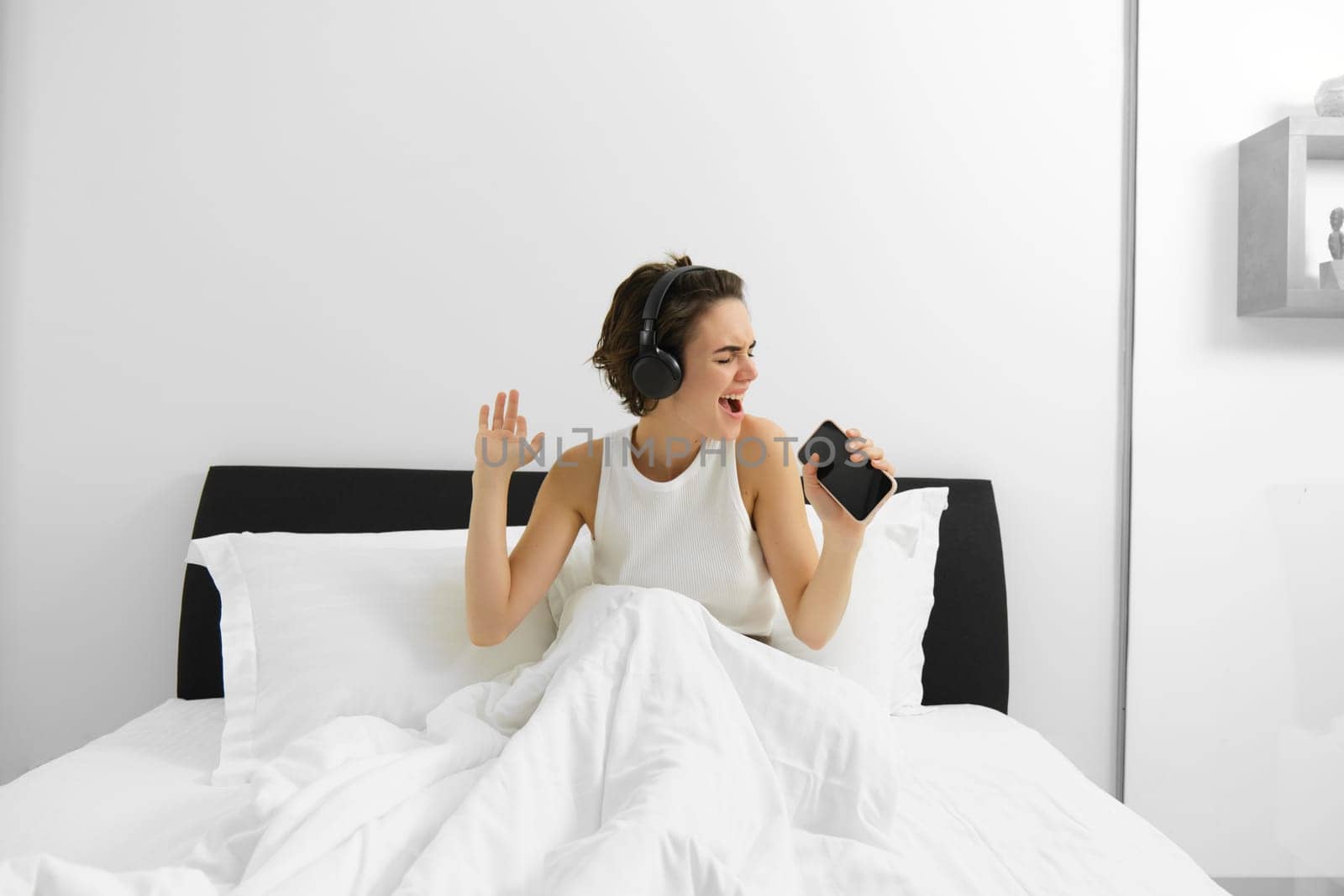 Carefree modern woman singing in her bedroom, into smartphone microphone, listening to music in wireless headphones by Benzoix