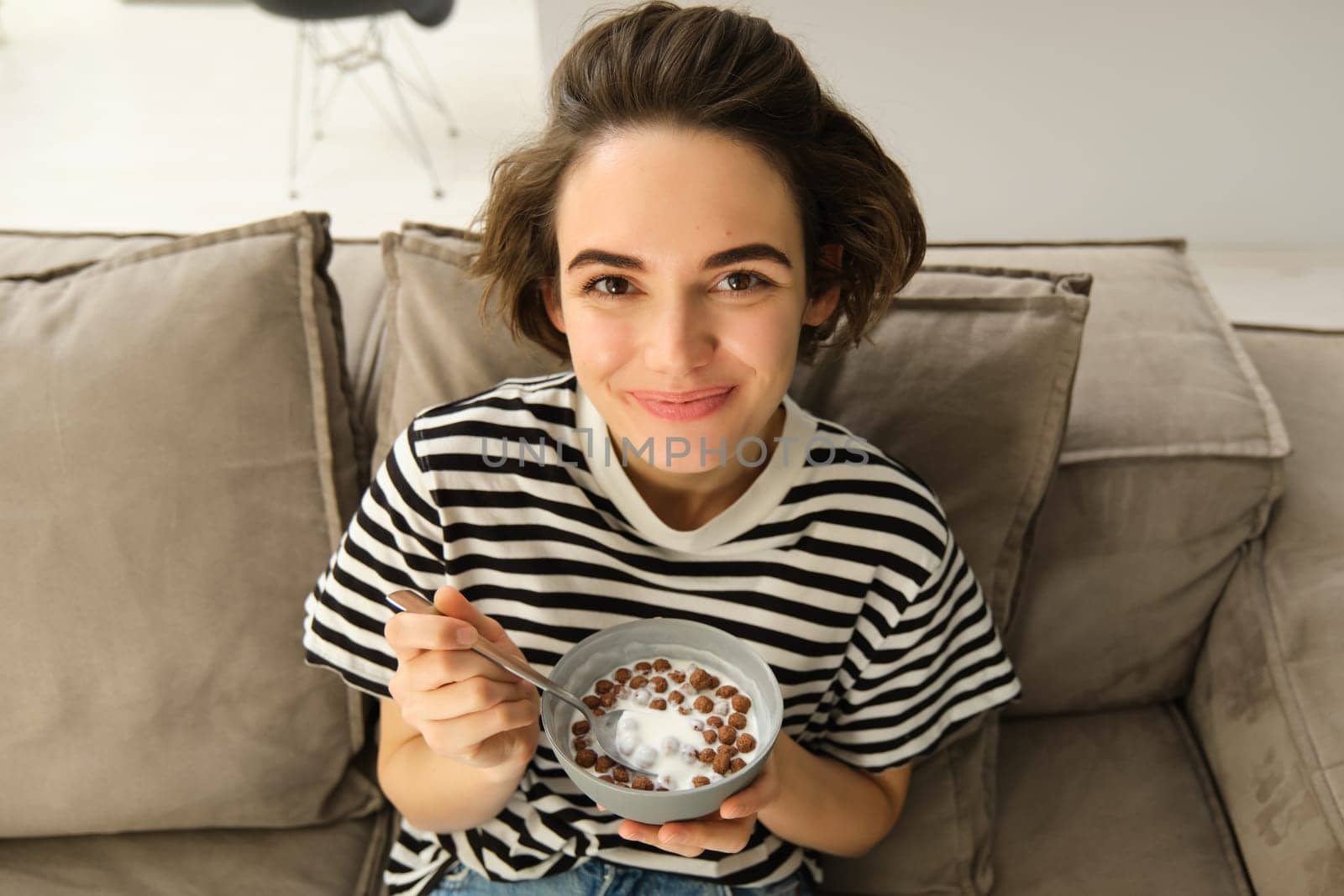 Close up of cute young female model, eating cereals with milk, enjoys her breakfast on sofa in living room, smiling and looking happy. People and food concept