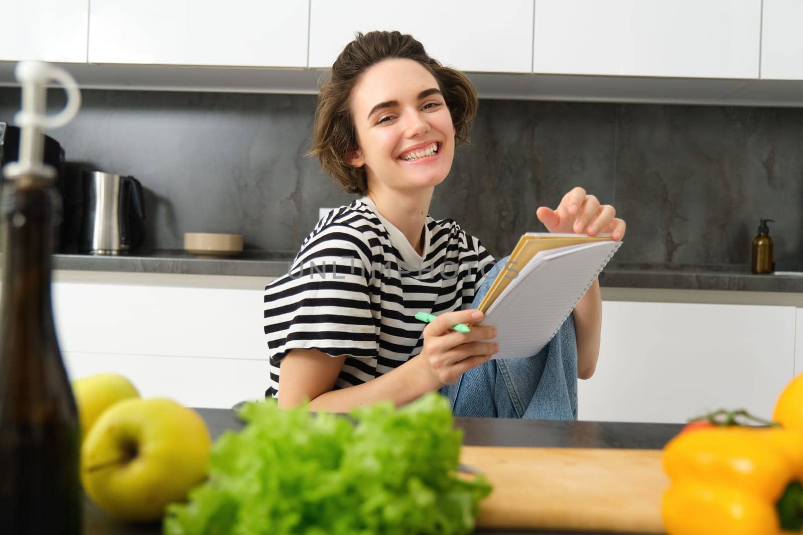 Portrait of young woman cooking, writing notes, grocery list in notebook, creating list of meals to cook through meal, sitting in kitchen near vegetables and chopping board by Benzoix