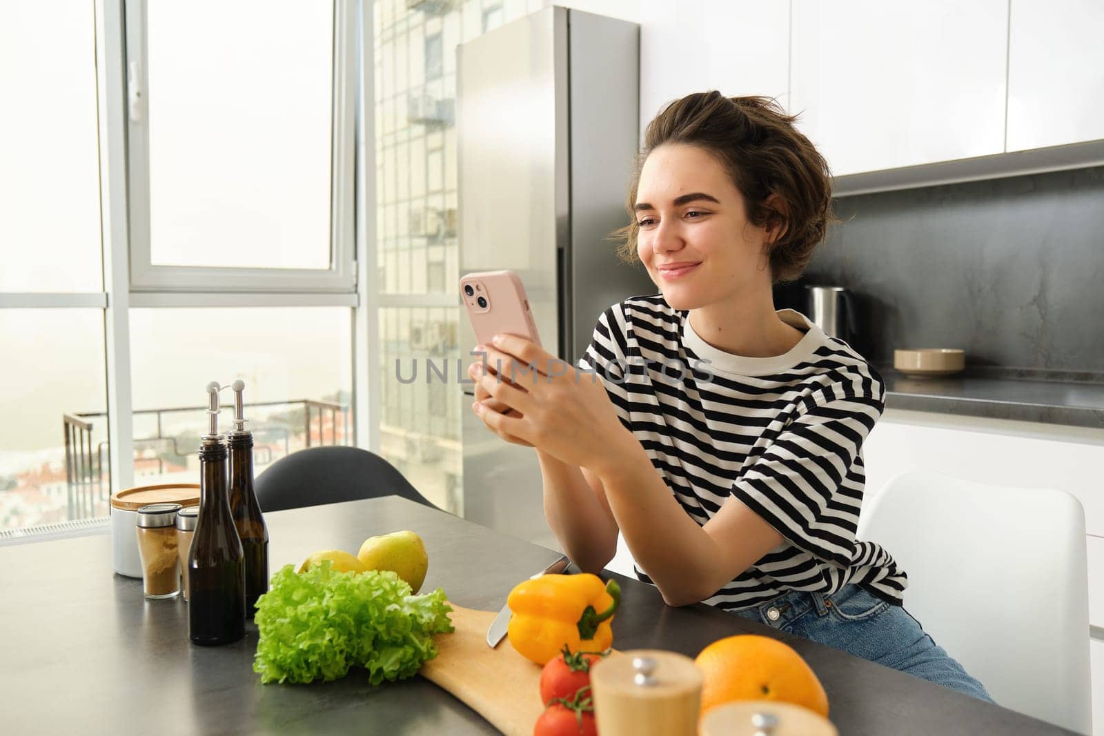 Portrait of cute smiling woman cooking salad, looking at smartphone, watching recipe, food preparation tutorial, chopping vegetables, making a meal in the kitchen. Healthy diet and lifestyle concept