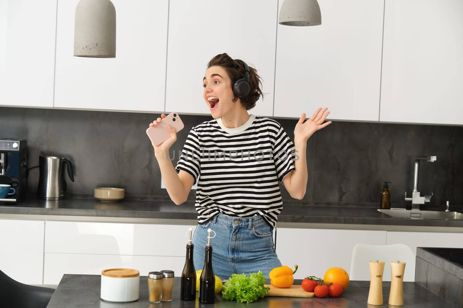 Portrait of carefree woman singing in smartphone mic, listening music in headphones while cooking breakfast, making a meal, chopping vegetables and dancing by Benzoix