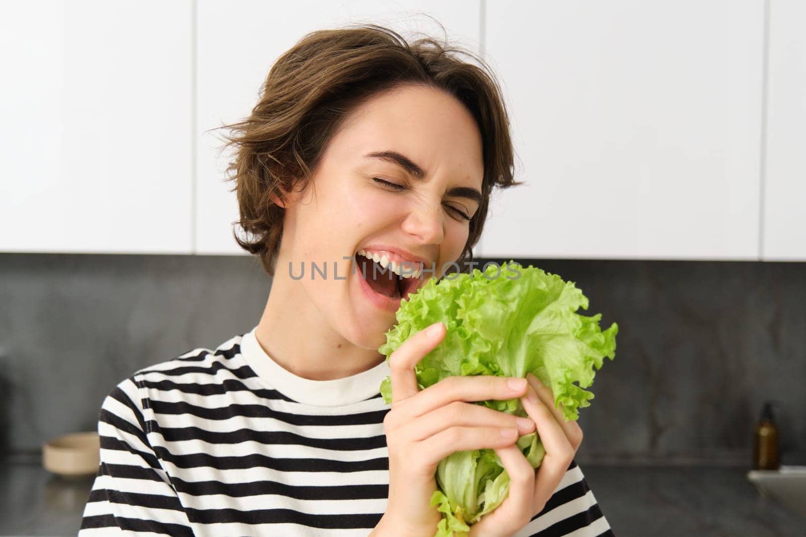 Portrait of happy and healthy young woman, following her diet, posing with lettuce leaf and smiling, cooking in the kitchen, vegetarian loves her vegetables by Benzoix