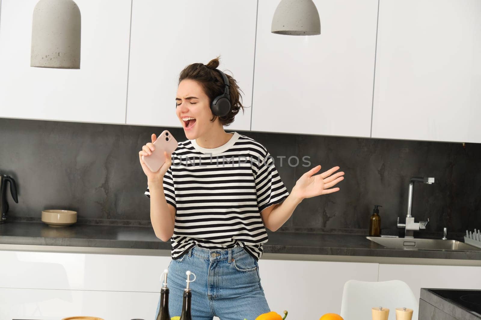 Portrait of carefree woman singing in smartphone mic, listening music in headphones while cooking breakfast, making a meal, chopping vegetables and dancing.