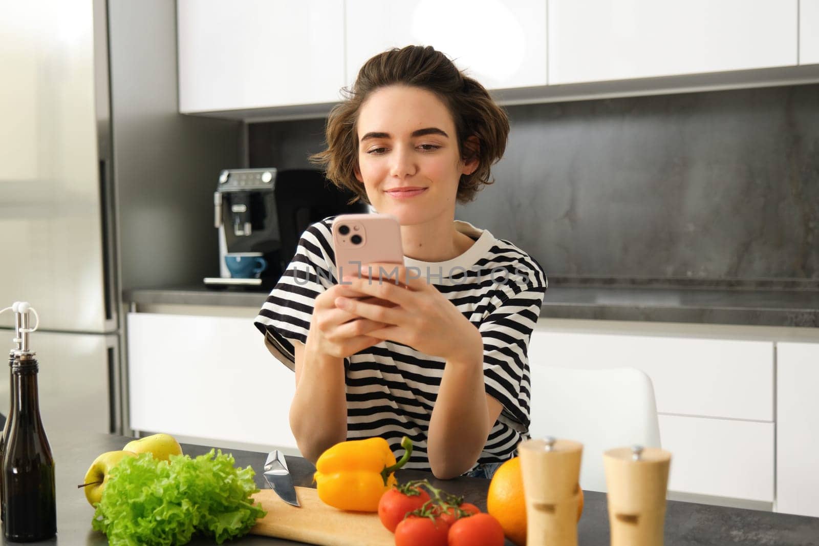 Portrait of smiling woman, holding smartphone, searching for healthy vegetarian recipe, standing near vegetables in the kitchen.