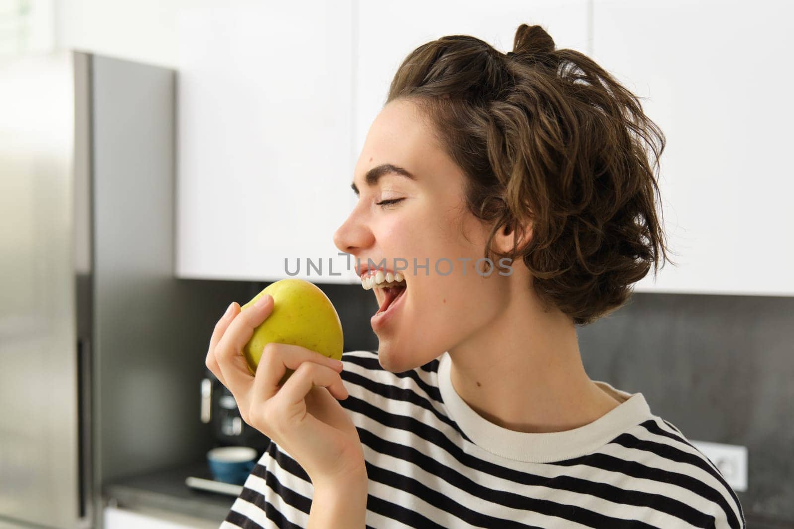 Close up portrait of young brunette woman biting an apple with pleasure, has pleased smile on her face, standing in the kitchen, having healthy snack for lunch, eating fruits by Benzoix