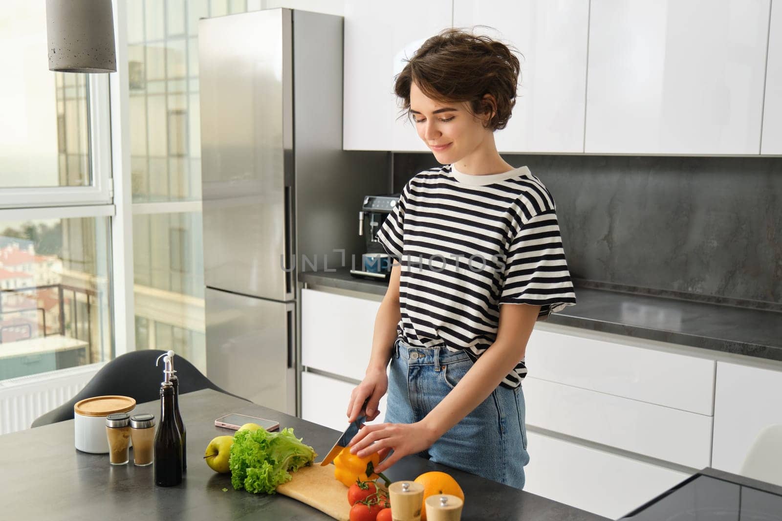 Portrait of smiling cute young woman making breakfast, chopping vegetables in the kitchen, preparing vegan meal.