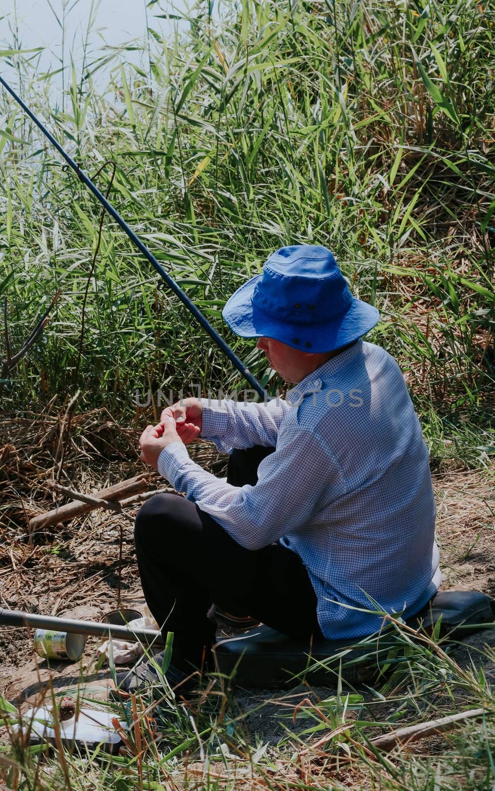One Caucasian young man in old clothes with a blue hat on his head puts a worm on a fishing rod hook while sitting on the shore of a lake in tall grass on a sunny summer day, close-up side view.