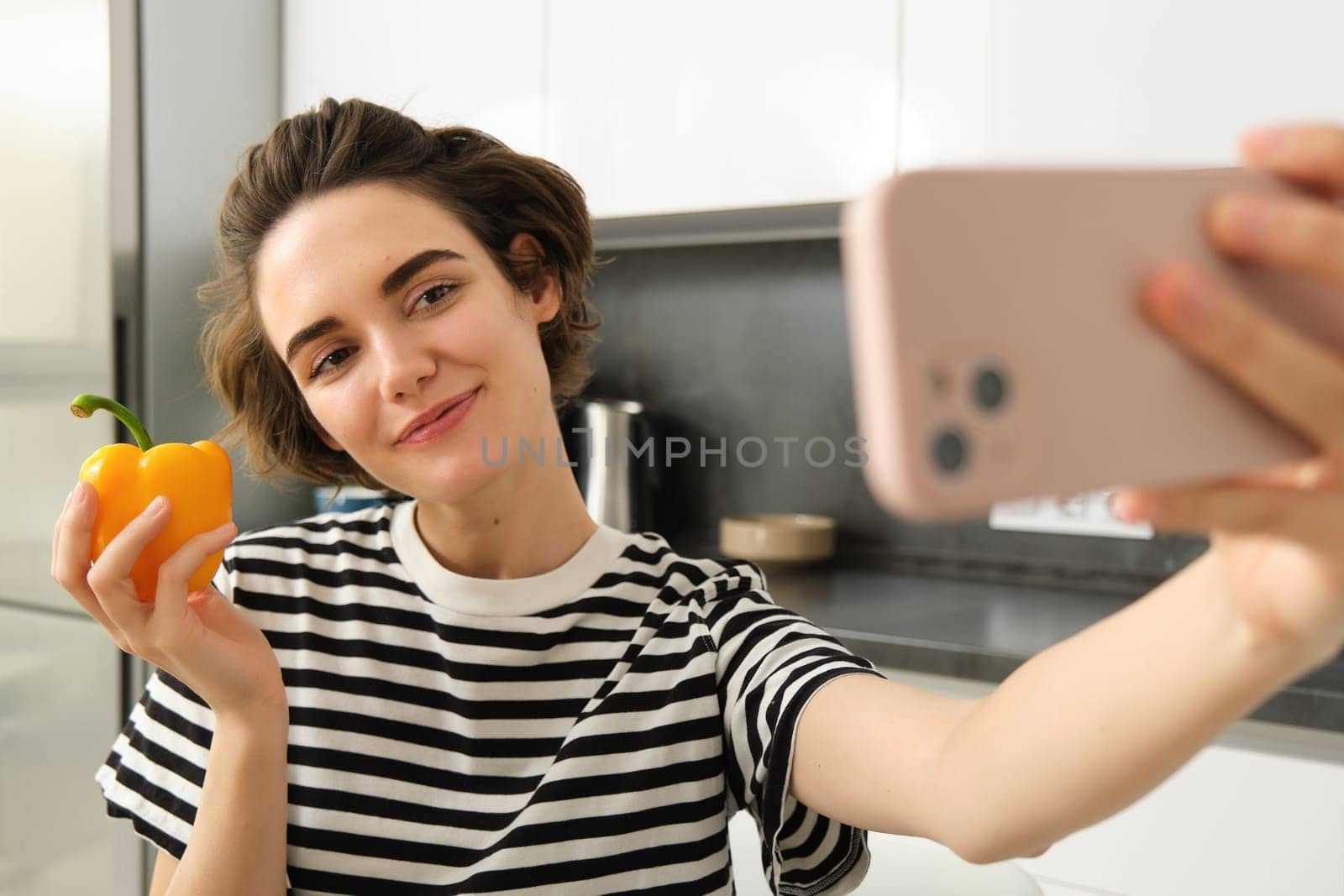 Cute and stylish lifestyle blogger, young woman taking selfie with fresh pepper, eating healthy vegetables, cooking salad in the kitchen and recording video on smartphone.