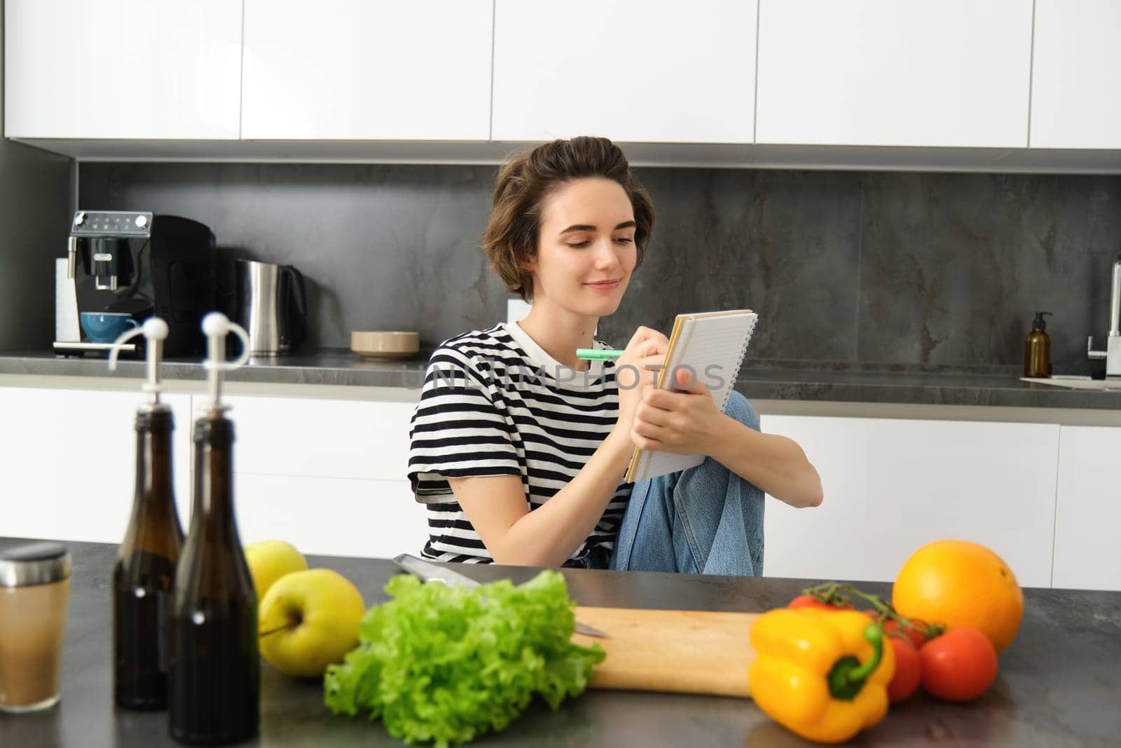 Portrait of beautiful modern woman, girl cooking with vegetables, holding notebook, writing down recipe, making notes, dinner plans, creating meal list for week, sitting in kitchen.